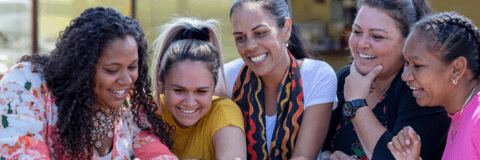 A photo of five women looking and smiling at a shared item that is not in the frame. 