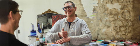 Image of a person signing while standing in front of many bowls of paint. A person in the foreground is smiling.