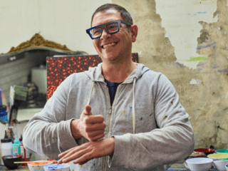 Image of a person signing while standing in front of many bowls of paint. A person in the foreground is smiling.