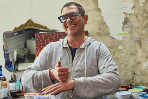 Image of a person signing while standing in front of many bowls of paint. A person in the foreground is smiling.